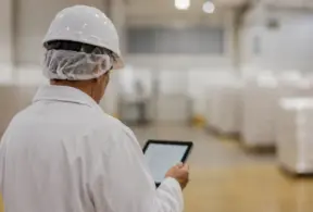Trabalhador de indústria de alimentos com capacete e máscara em frigorífico, usando tablet para acompanhar processos na linha de produção; foco em monitoramento socioambiental
