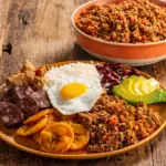 Prato com cortes de carne bovina acompanhados de ovo frito, feijão e banana da terra, ao lado de tigela com carne moída sobre mesa de madeira