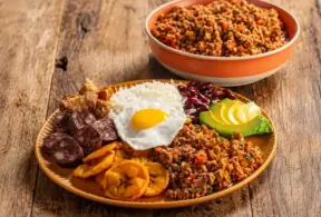 Prato com cortes de carne bovina acompanhados de ovo frito, feijão e banana da terra, ao lado de tigela com carne moída sobre mesa de madeira