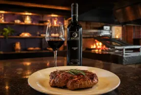 carne e vinho servidos em ambiente gourmet: prato com bife grelhado e taça com vinho tinto ao lado de uma garrafa em cenário de churrasqueira e luz aconchegante