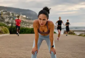 Mulher correndo em uma estrada costeira com outras pessoas ao fundo, praticando atividade física ao ar livre ao pôr do sol em clima esportivo