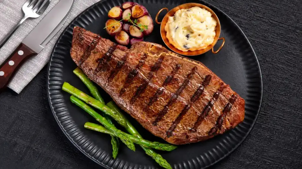nomes diferentes de carne: Short rib grelhado em prato preto com aspargos verdes e acompanhamento cremoso em tigela pequena, com porções de linguiça e batatas na lateral; refeição pronta.