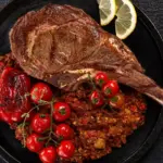 nomes diferentes de carne: Tomahawk grelhado em uma panela com batatas ao estilo crocante, acompanhado de tomates-cereja e fatias de limão, em fundo escuro de madeira