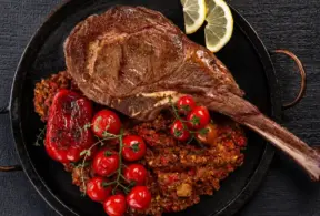 nomes diferentes de carne: Tomahawk grelhado em uma panela com batatas ao estilo crocante, acompanhado de tomates-cereja e fatias de limão, em fundo escuro de madeira