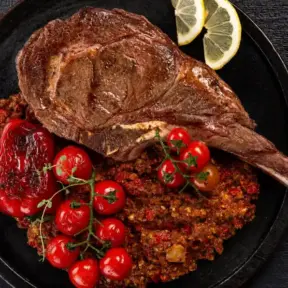 nomes diferentes de carne: Tomahawk grelhado em uma panela com batatas ao estilo crocante, acompanhado de tomates-cereja e fatias de limão, em fundo escuro de madeira