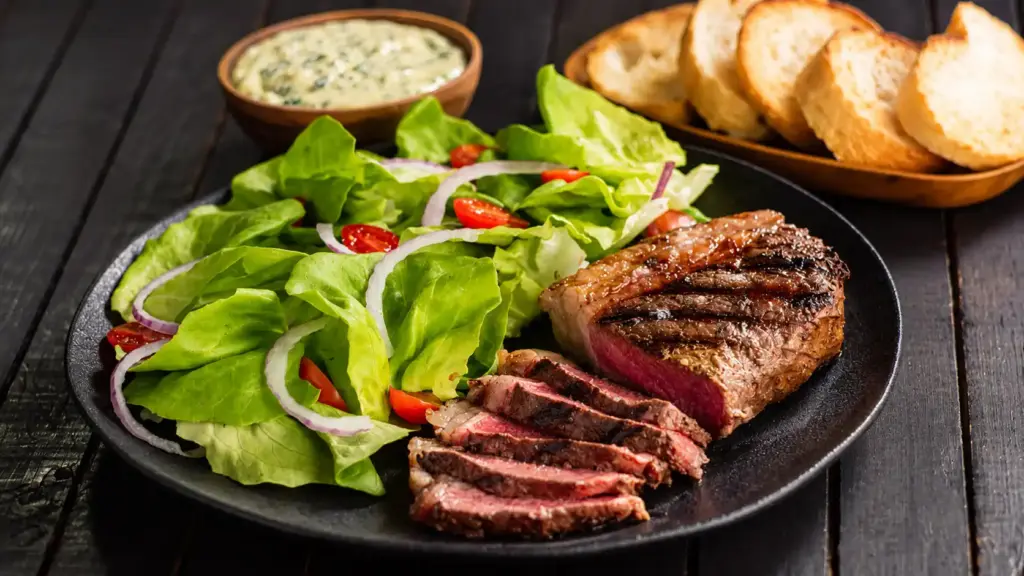 Segurança alimentar: prato com fatias de carne grelhada, salada de folhas verdes e tomate-cereja, acompanhado de pães em fundo escuro
