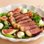 Salada gourmet em prato branco com fatias de carne bovina, folhas verdes, rúcula, figo fatiado, ovos de codorna e molhos, em mesa clara. Ciência da carne aplicada à preparação de pratos com ingredientes frescos.