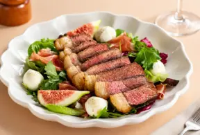 Salada gourmet em prato branco com fatias de carne bovina, folhas verdes, rúcula, figo fatiado, ovos de codorna e molhos, em mesa clara. Ciência da carne aplicada à preparação de pratos com ingredientes frescos.