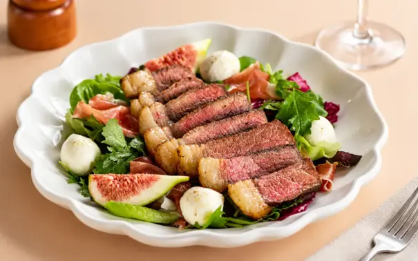 Salada gourmet em prato branco com fatias de carne bovina, folhas verdes, rúcula, figo fatiado, ovos de codorna e molhos, em mesa clara. Ciência da carne aplicada à preparação de pratos com ingredientes frescos.