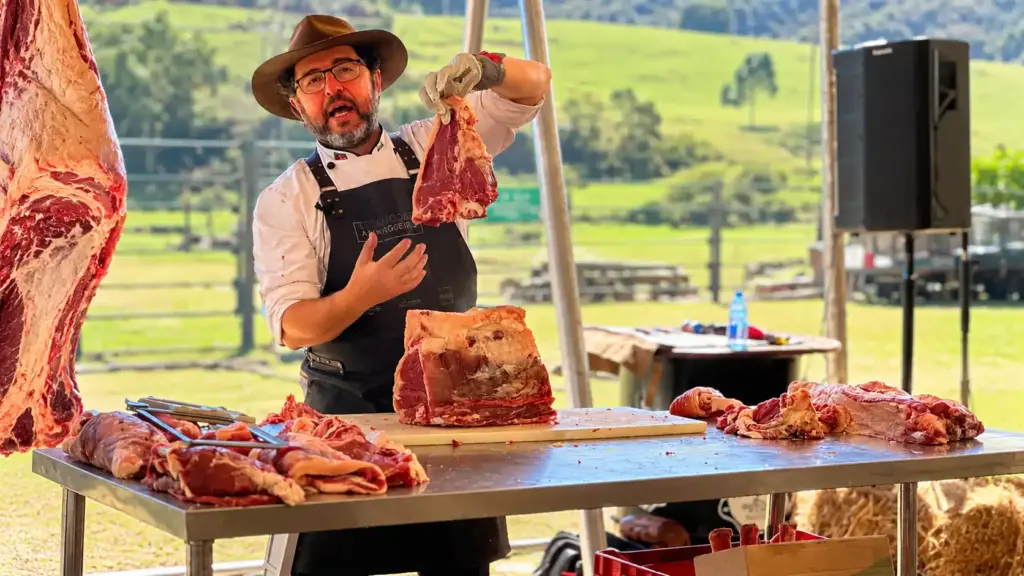 Marcelo Bolinha, açougueiro do churrasco, segura um grande corte de carne em um churrasco ao ar livre, com mesas e pedaços de carne preparados no fundo.