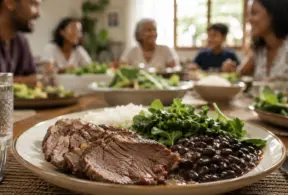 Close-up de um prato com fatias de carne bovina, feijão preto, arroz e verduras — deliciosos alimentos ricos em ferro — sobre uma mesa. Ao fundo, quatro pessoas estão sentadas juntas, compartilhando uma refeição, sorrindo e conversando.