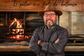 Marcelo Bolinha, açougueiro e especialista em churrasco, em frente a um forno com chamas e grelha, com uniforme e chapéu, braços cruzados.