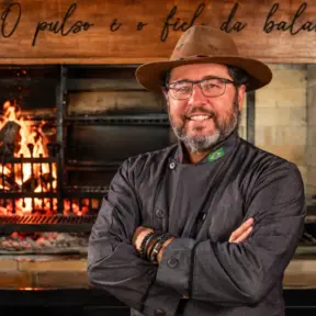 Marcelo Bolinha, açougueiro e especialista em churrasco, em frente a um forno com chamas e grelha, com uniforme e chapéu, braços cruzados.