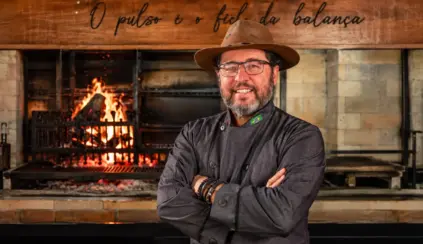 Marcelo Bolinha, açougueiro e especialista em churrasco, em frente a um forno com chamas e grelha, com uniforme e chapéu, braços cruzados.