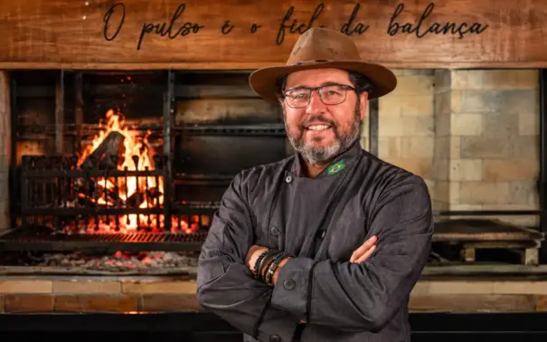 Marcelo Bolinha, açougueiro e especialista em churrasco, em frente a um forno com chamas e grelha, com uniforme e chapéu, braços cruzados.