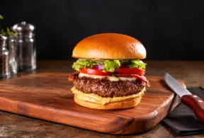 hambúrguer caseiro com carne grelhada, queijo e fatias de tomate sobre pão brioche, servido em tábua de madeira com faca ao lado e temperos na bancada