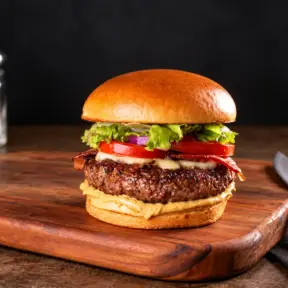 hambúrguer caseiro com carne grelhada, queijo e fatias de tomate sobre pão brioche, servido em tábua de madeira com faca ao lado e temperos na bancada