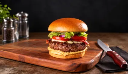 hambúrguer caseiro com carne grelhada, queijo e fatias de tomate sobre pão brioche, servido em tábua de madeira com faca ao lado e temperos na bancada