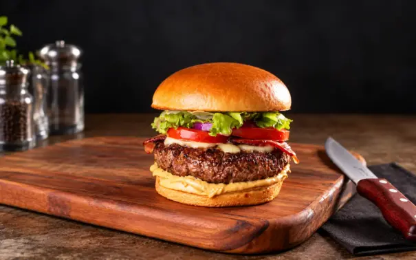 hambúrguer caseiro com carne grelhada, queijo e fatias de tomate sobre pão brioche, servido em tábua de madeira com faca ao lado e temperos na bancada