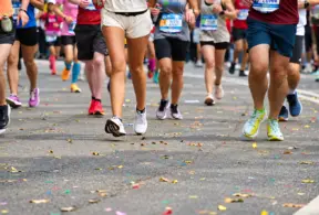 Um grupo de corredores, com números de inscrição bem visíveis, corre por uma rua coberta de confete colorido durante uma corrida de rua ou maratona. Apenas suas pernas e pés estão visíveis, demonstrando a dedicação e a energia alimentadas pela proteína para corredores.