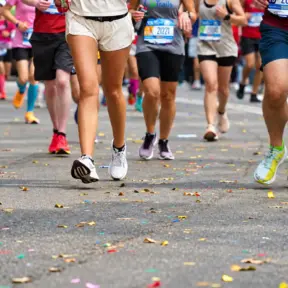 Um grupo de corredores, com números de inscrição bem visíveis, corre por uma rua coberta de confete colorido durante uma corrida de rua ou maratona. Apenas suas pernas e pés estão visíveis, demonstrando a dedicação e a energia alimentadas pela proteína para corredores.