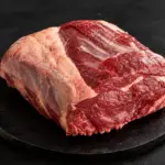 Peça de carne bovina crua em corte com gordura aparente sobre uma base preta de pedra, pronta para preparo culinário; carne de segunda em destaque.