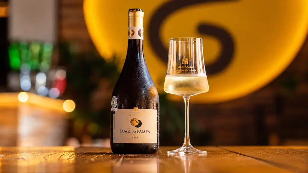 Uma garrafa de Luar do Pampa Chardonnay, um excelente vinho para churrasco, ao lado de uma taça de vinho cheia sobre uma mesa de madeira.