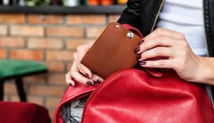 Mulher segura uma bolsa vermelha com uma carteira marrom acoplada, em ambiente externo com parede de tijolos ao fundo. dia nacional da pecuária