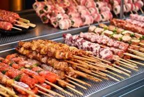 Churrasco na China com espetinhos grelhados em uma chapa industrial, com legumes e ervas na composição dos pratos.
