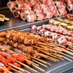 Churrasco na China com espetinhos grelhados em uma chapa industrial, com legumes e ervas na composição dos pratos.