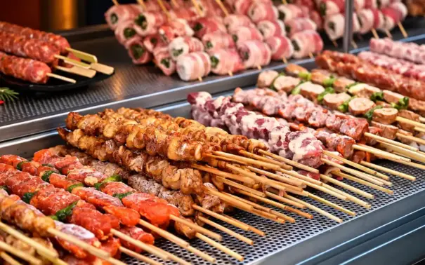 Churrasco na China com espetinhos grelhados em uma chapa industrial, com legumes e ervas na composição dos pratos.