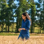 Mulher caminhando em uma plantação em meio a árvores no campo, com expressão serena, representando pecuária baixa emissão e sustentabilidade no meio rural ao ar livre.