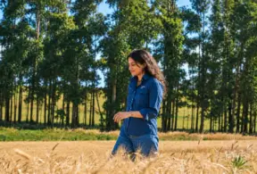 Mulher caminhando em uma plantação em meio a árvores no campo, com expressão serena, representando pecuária baixa emissão e sustentabilidade no meio rural ao ar livre.