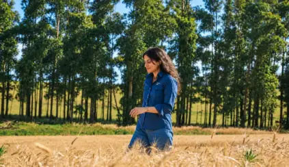 Mulher caminhando em uma plantação em meio a árvores no campo, com expressão serena, representando pecuária baixa emissão e sustentabilidade no meio rural ao ar livre.