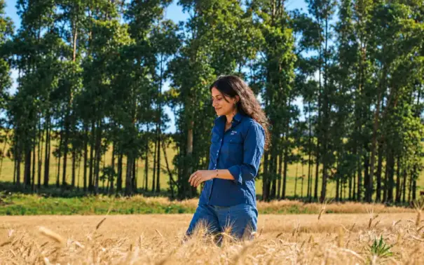 Mulher caminhando em uma plantação em meio a árvores no campo, com expressão serena, representando pecuária baixa emissão e sustentabilidade no meio rural ao ar livre.