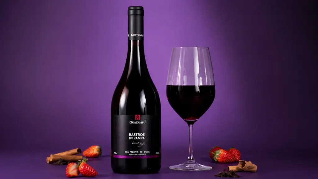 Uma garrafa de Guatambu Rastros do Pampa Tannat, um vinho perfeito para churrasco, ao lado de uma taça de vinho tinto, rodeada por morangos e paus de canela contra um fundo roxo.
