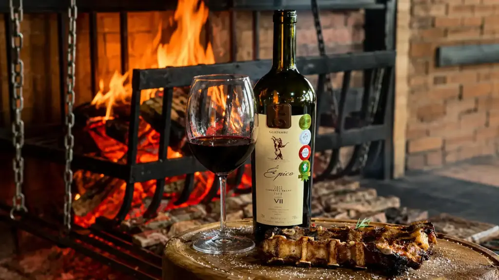 Uma taça de vinho tinto, uma garrafa de Guatambu Épico Cabernet Franc e uma porção de costelas servida em uma tábua de madeira formam a combinação perfeita de vinho para churrasco em frente à lareira.