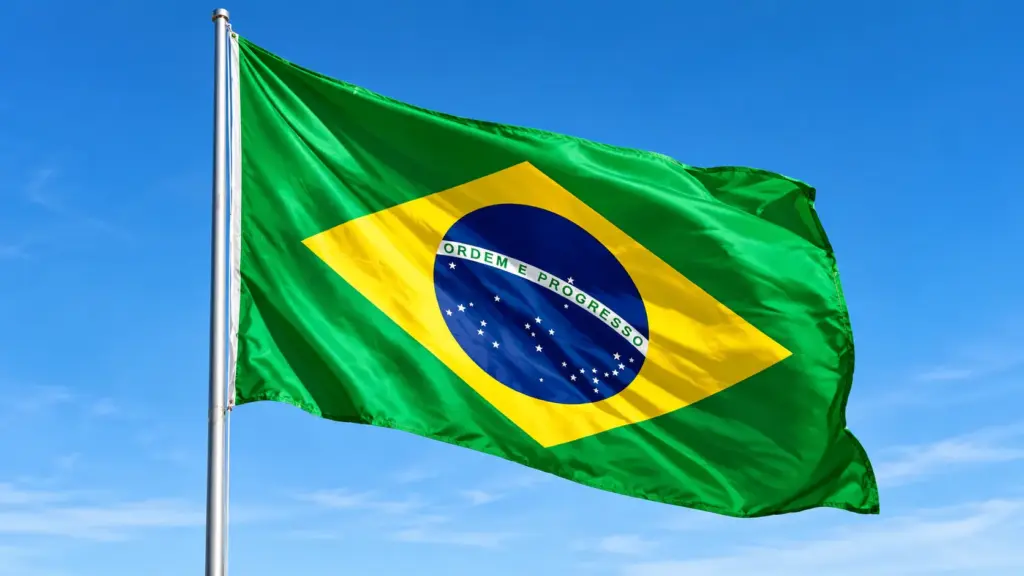 proteína: Bandeira do Brasil ao vento, com o lema “Ordem e Progresso” e o círculo azul com estrelas sobre fundo verde e amarelo, no céu azul.