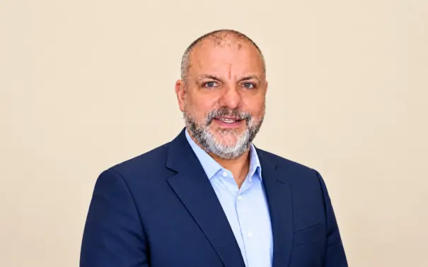 Pedro Loyola usando terno azul-marinho e camisa social clara, em fundo liso claro, sorrindo para a câmera. Foto corporativa.
