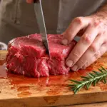Homem corta carne em um pedaço de carne na tábua de madeira, com faca e temperos como alecrim e alho ao lado, em preparação de receita de carne solta água.