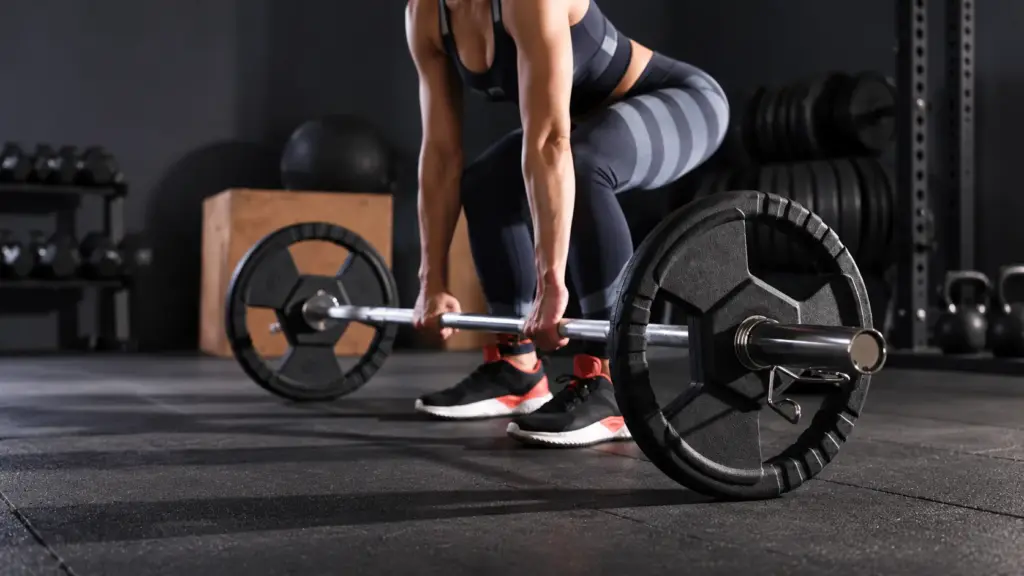 Uma pessoa vestida com roupa esportiva, interessada em ganhar massa muscular, se preparando para levantar uma barra do chão da academia.