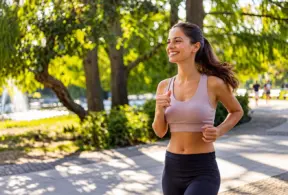 Mulher sorridente correndo ao ar livre em um parque, vestida com roupas esportivas e sob o sol da manhã, praticando corrida para perder peso.