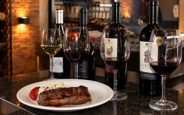 Mesa posta com taças de vinho tinto e um prato com carne grelhada, pimentão e queijo, em ambiente com lareira e destaque para vinho para churrasco
